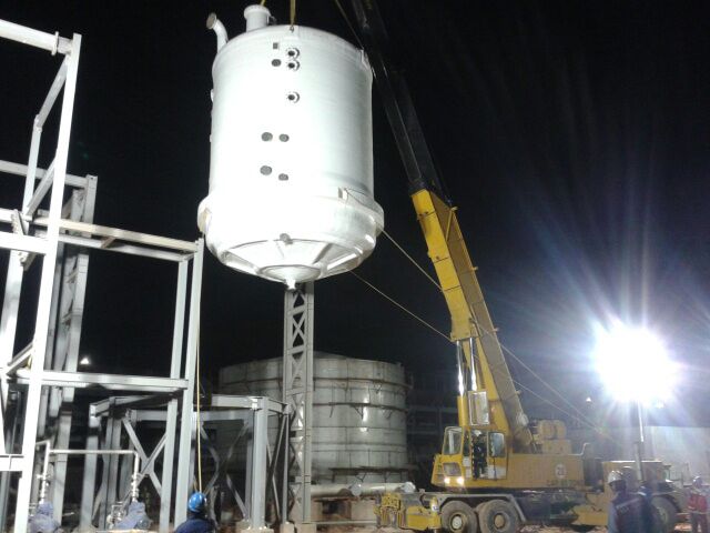 Large white FRP tank with conical bottom being lifted by crane during nighttime installation at a mining plant