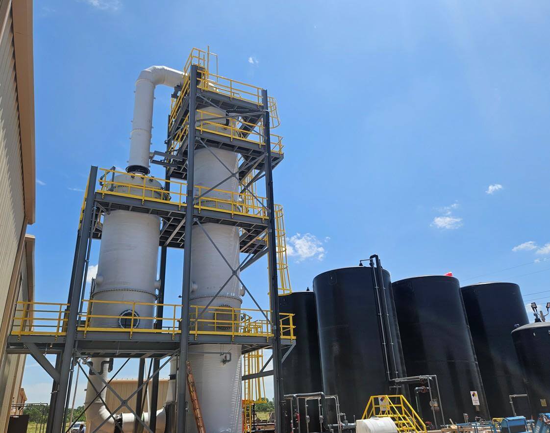 White FRP scrubber towers and ductwork on a steel access structure with yellow handrails, next to black storage tanks at an industrial plant